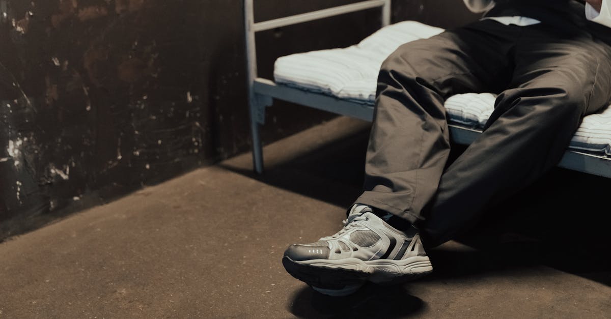 man in casual attire sits on a simple bed in an industrial setting holding a paper